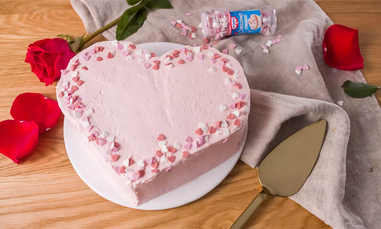 Pastel en forma de corazón con crema suave rosada, borde decorado con corazones de azúcar, sobre plato blanco y mantel beige sobre madera