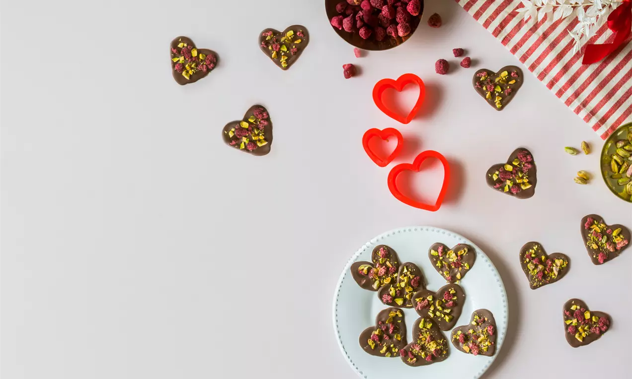 Corazones de chocolate oscuro decorados con pistachos y frambuesas secas en un plato blanco, rodeados de moldes rojos y frutos