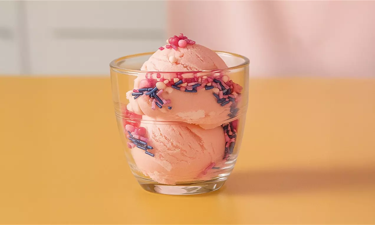 Bolas de helado de fresa rosado suave servidas en vaso transparente decoradas con chispas coloridas y perlas rosas sobre mesa amarilla