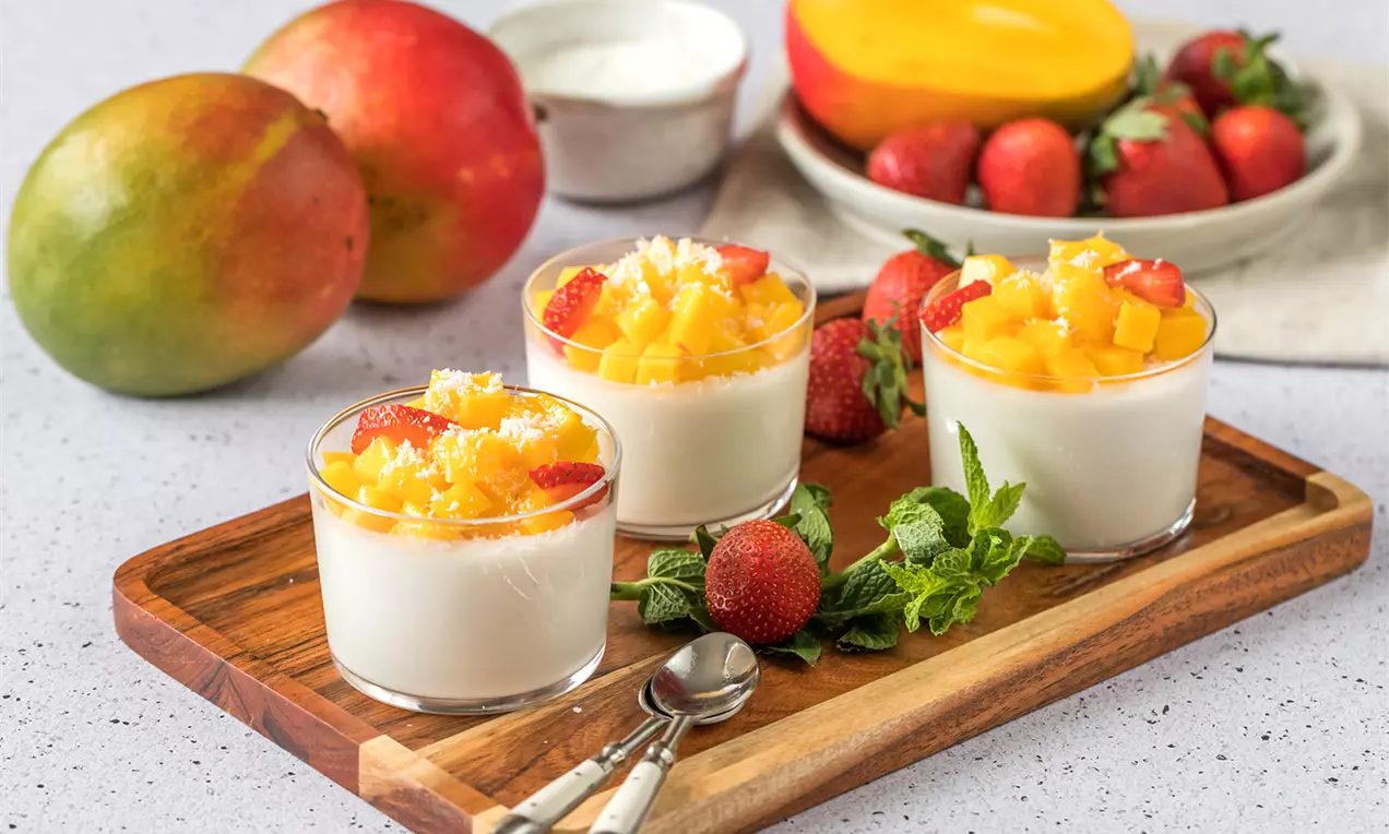 Vasitos de crema blanca suave coronados con cubos de mango amarillo brillante, fresas cortadas y coco rallado sobre una tabla de madera