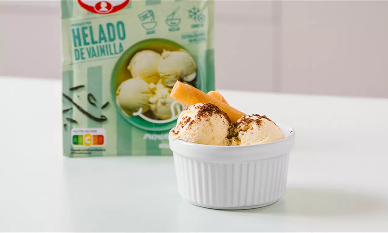 Helado de vainilla cremoso y suave en una cazuela blanca, decorado con barquillos dorados y espolvoreado con cacao sobre una mesa blanca