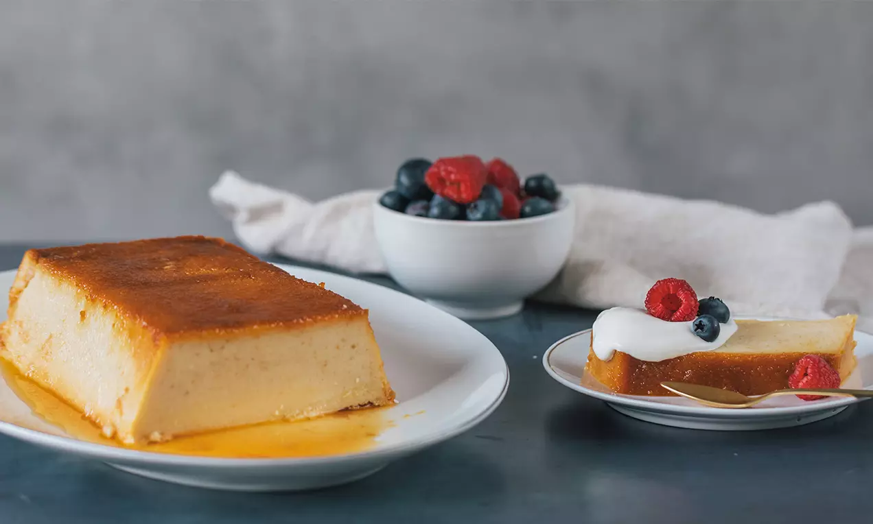 Rebanada de pudín de pan dorado y cremoso con textura suave sobre plato blanco, decorada con nata, frambuesas y arándanos