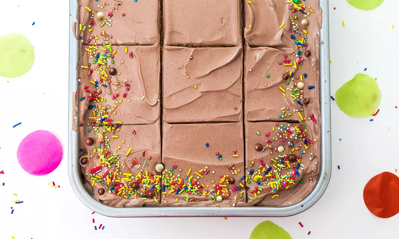Tarta rectangular de bizcocho con cubierta cremosa de chocolate partido en porciones, decorada con chispas de colores sobre bandeja metálica