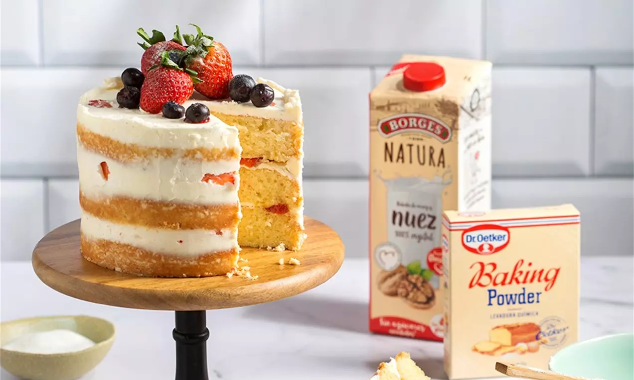 Pastel de capas doradas y esponjosas con relleno blanco, decorado con fresas rojas y arándanos sobre una base de madera