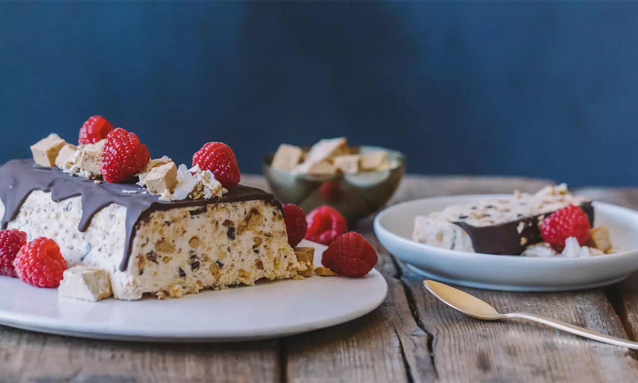 Semifrío cremoso beige con trozos de almendra, cubierto de chocolate brillante, frambuesas frescas y dados de turrón sobre plato blanco