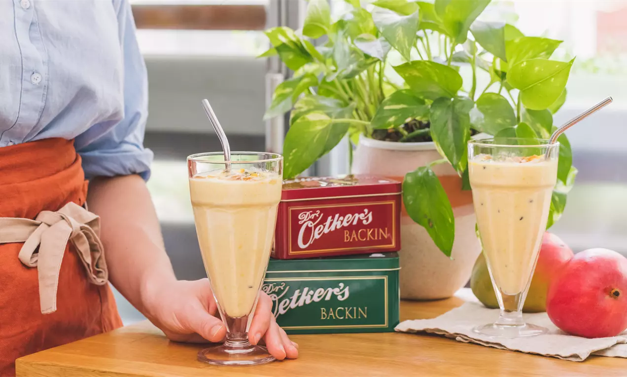 Batido amarillo cremoso en vaso alto con pajita metálica, junto a cajas de colores, frutas frescas y una planta verde sobre madera clara