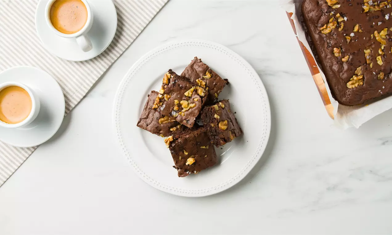 Cuadrados de brownie marrón oscuro con superficie ligeramente crujiente, trozos de nuez dorada encima y servidos en un plato blanco